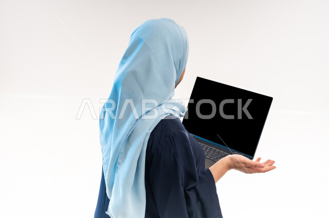 اجراء مقابلات عمل اونلاين، دمج التقنية والتكنولوجيا الحديثة في الحياة العملية، بورتريه مقرب من الخلف لامرأة عربية خليجية عمانية ترتدي العباءة تحمل بيدها عنصر ما، شابة سعودية ترفع كفها بإيماءات عدم المعرفة، عرض شاشة الحاسوب المحمول السوداء، خلفية بيضاء