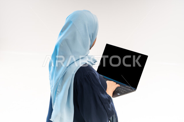 دمج التقنية والتكنولوجيا الحديثة في الحياة اليومية، بورتريه مقرب من الخلف لامرأة عربية خليجية عمانية ترتدي العباءة تقوم بالنقر على لوحة مفاتيح اللابتوب، تصفح مواقع ووسائل التواصل الاجتماعي، شابة سعودية تعمل على جهاز الحاسوب المحمول، خلفية بيضاء