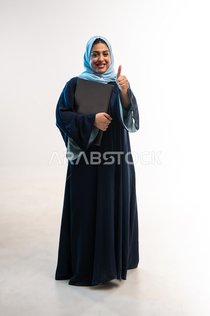 مهن ووظائف نسائية, ، دمج التقنية والتكنولوجيا الحديثة في الحياة العملية، بورتريه لامرأة عربية خليجية اماراتية ترتدي العباءة تحمل بيدها جهاز الحاسوب المحمول، شابة سعودية ترفع اصبع الابهام بإيماءات الاعجاب، صورة لكامل الجسم، خلفية بيضاء
