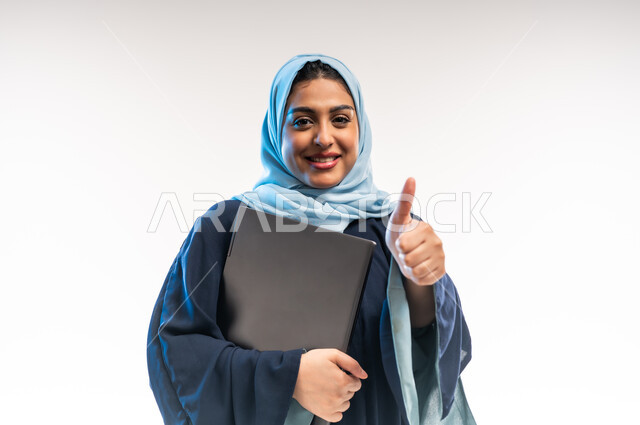 رفع اصبع الابهام بإيماءات الاعجاب، مهن ووظائف نسائية, دمج التقنية والتكنولوجيا الحديثة في الحياة العملية، بورتريه مقرب لامرأة عربية خليجية اماراتية ترتدي العباءة تحمل بيدها جهاز الحاسوب المحمول، شابة سعودية تنظر الى الكاميرا بتعابير الثقة بالنفس، خلفية بيضاء