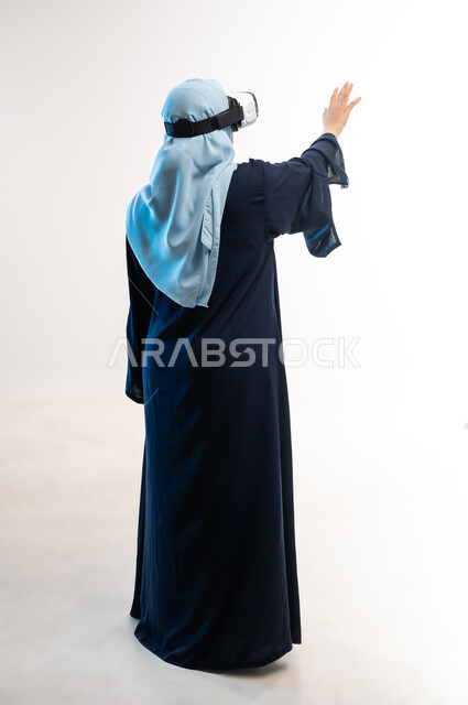 الاندماج ومحاكاة العالم الخيالي، المتعة والتسلية بالمشاهدة عبر نظارة الواقع الافتراضي، بورتريه من الخلف لإمرأة عربية خليجية عمانية محجبة تنظر من خلال نظارات ثلاثية الأبعاد، شابة سعودية ترفع يديها للاعلى لمحاولة لمس شيء ما،صورة لكامل الجسم، خلفيه بيضاء
