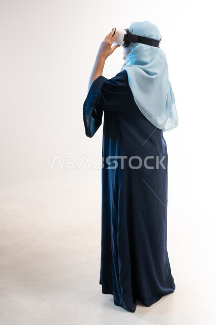 الاندماج ومحاكاة العالم الخيالي، المتعة والتسلية بالمشاهدة عبر نظارة الواقع الافتراضي، بورتريه من الخلف لإمرأة عربية خليجية عمانية محجبة تنظر من خلال نظارات ثلاثية الأبعاد، شابة سعودية تقوم باستخدام التقنيات الحديثة في الترفيه، صورة لكامل الجسم، خلفيه بيضاء