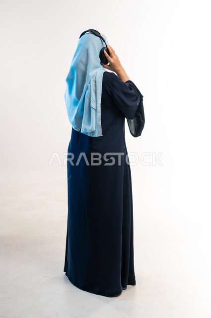 مفهوم الإصغاء والإستماع، قضاء اوقات ممتعة ومسلية، إستخدام تقنيات الهيدفون، بورتريه من الخلف لامرأة عربية خليجية عمانية محجبة تضع سماعات الرأس الصوتية تستمع إلى الموسيقى، شابة سعودية تمسك بالسماعة، صورة لكامل الجسم، خلفية بيضاء