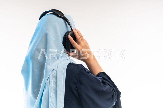 قضاء اوقات ممتعة ومسلية، مفهوم الإصغاء والإستماع، إستخدام تقنيات الهيدفون، بورتريه مقرب من الخلف لامرأة عربية خليجية عمانية محجبة تضع سماعات الرأس الصوتية تستمع إلى الموسيقى، شابة سعودية تمسك بالسماعة، خلفية بيضاء