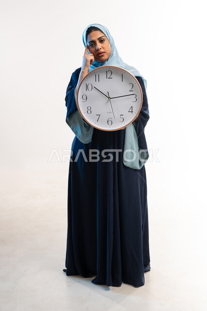Constant reminders and alerts, importance of keeping time, completing tasks on time, portrait of a veiled Omani Gulf Arab woman wearing a blue abaya holding a wall clock in her hand, a young Saudi woman resting it on her chin with boredom gestures, full body image, white background
