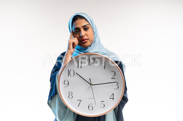 أهمية الحفاظ على الأوقات, التذكيرات والتنبيهات الثابتة, إنجاز المهام في الموعد المحدد, بورتريه مقرب لامرأة عربية خليجية عمانية محجبة ترتدي عباءة زرقاء تمسك بيدها ساعة حائط ، شابة سعودية تضعيدها على ذقنها بايماءات الملل, خلفية بيضاء