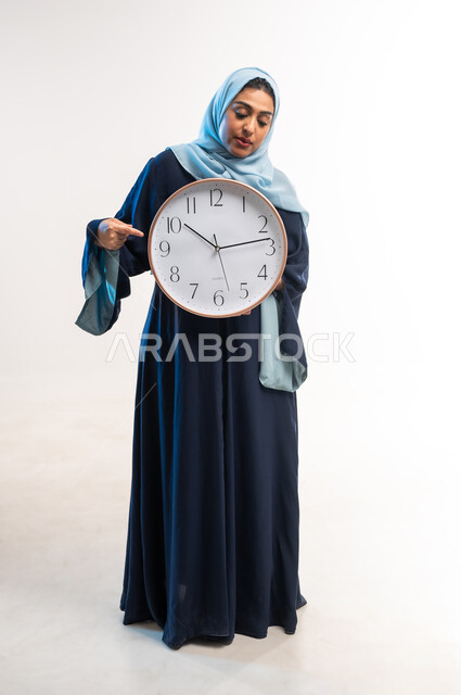 إنجاز المهام في الموعد المحدد, التذكيرات والتنبيهات الثابتة, أهمية الحفاظ على الأوقات, بورتريه لامرأة عربية خليجية عمانية محجبة ترتدي عباءة زرقاء تمسك بيدها ساعة حائط ، شابة سعودية تشير بإصبع السبابة إلى وقت معين, الجسم بالطول الكامل, خلفية بيضاء