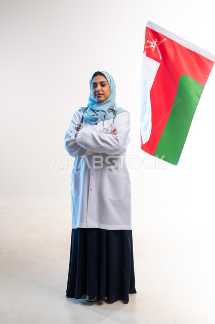 إحياء ذكرى استقال الدولة 1650م، رفع علم سلطنة عمان، اليوم الوطني العماني 18 نوفمبر, العمل في المجال الطبي, بورتريه لطبيبة عربية خليجية عمانية ترتدي المعطف الطبي والسماعة تقف بايدي متقاطعة تنظر الى الكاميرا بايماءات الفخر، صورة لكامل الجسم، خلفية بيضاء