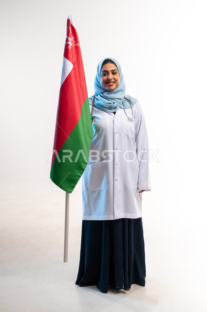 اليوم الوطني العماني 18 نوفمبر, إحياء ذكرى استقال الدولة 1650م، مفهوم الطب والرعاية الصحية, العمل في المراكز والمستشفيات, بورتريه لطبيبة عربية خليجية عمانية ترتدي المعطف الطبي وسماعة الفحص تحمل بيدها علم سلطنة عمان، صورة لكامل الجسم، خلفية بيضاء