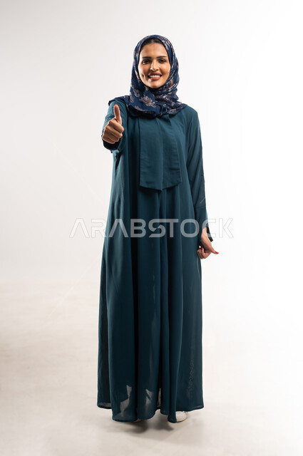 تعابير الموافقة والقبول, سيدة سعودية تنظر الى الكاميرا بإيماءات السرور الرضى والاعجاب، بورتريه لامرأة عربيه عمانية مبتسمة محجبة ترتدي العباءه تشير باصبع الابهام الى الاعلى بعلامه الامتياز، الوقوف باستقامة وثبات،الطول الكامل للجسم، خلفية بيضاء