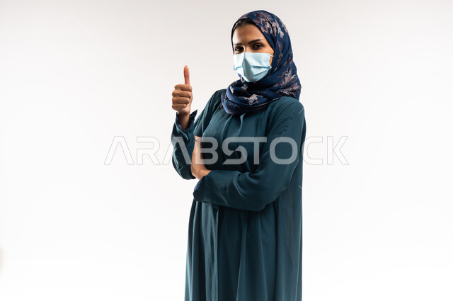 اتخاذ التدابير للوقاية من فيروس كورونا، الاهتمام بالاناقة والمظهر الخارجي، سيدة سعودية تنظر الى الكاميرا وتضع الكمامة بتعابير الموافقة والقبول, بورتريه لامرأة عربيه عمانية محجبة ترتدي العباءه ترفع اصبع الابهام بايماءات الرضا والموافقة، خلفية بيضاء
