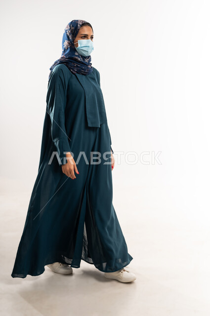 ايماءات الذهاب إلى مكان معين، بورتريه من الجانب لإمرأة عربية خليجية عمانية محجبة ترتدي العباءة تنظر إلى شيء ما، التقدم إلى الأمام بخطوات ثابتة، سيدة سعودية تضع الكمامة الطبية لاتخاذ الإجراءات الوقائية، مفهوم الأناقة والأنوثة, الطول الكامل للجسم، خلفية بيضاء