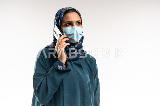 إجراء مكالمة صوتية عبر الجوال، دمج التقنيات الحديثة بالحياة اليومية، بورتريه مقرب لامرأة عربية خليجية عمانية ترتدي العباءة والكمامة لاتخاذ إجراءات الوقاية من كورونا، شابة سعودية تحمل بيدها الهاتف المحمول، التواصل مع الأهل والأصدقاء، خلفية بيضاء