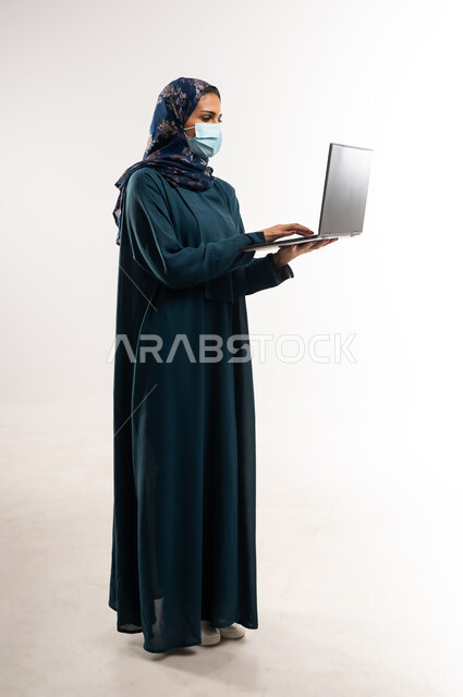 دمج التقنية والتكنولوجيا الحديثة في الحياة اليومية، بورتريه من الجانب لامرأة عربية خليجية عمانية ترتدي العباءة تقوم بالنقر على لوحة مفاتيح اللابتوب، تصفح مواقع ووسائل التواصل الاجتماعي، شابة سعودية تعمل على جهاز الحاسوب المحمول، الطول الكامل للجسم، خلفية بيضاء