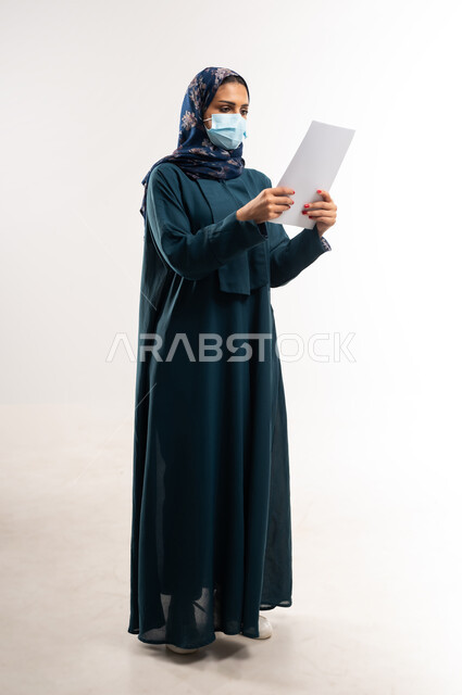 الحفاظ على السلامة من كوفيد 19، لوحات توعوية لتجنب إنتشار العدوى، اتباع الاجراءات الاحترازية للوقاية من فايروس كورونا، بورتريه لإمرأة عربية خلجية عمانية محجبة ترتدي العباءة وتضع الكمامة، سيدة سعودية تمسك أوراق بيضاء فارغة, الجسم بالطول الكامل، خلفية بيضاء