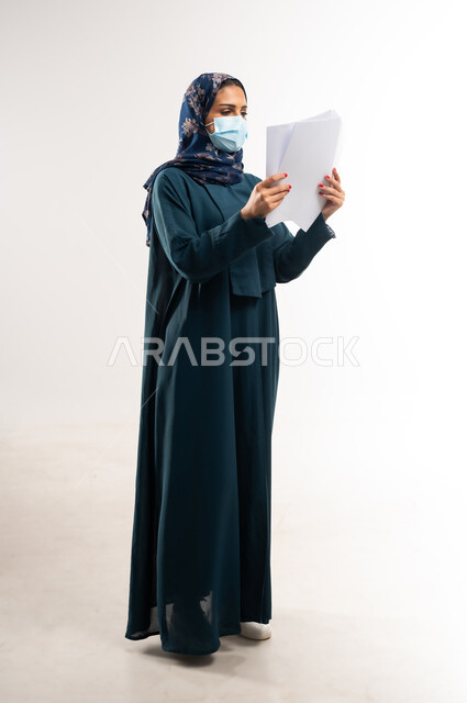 الحفاظ على السلامة من كوفيد 19، لوحات توعوية لتجنب إنتشار العدوى، اتباع الاجراءات الاحترازية للوقاية من فايروس كورونا، بورتريه لإمرأة عربية خلجية عمانية محجبة ترتدي العباءة وتضع الكمامة، سيدة سعودية تمسك أوراق بيضاء فارغة, الجسم بالطول الكامل، خلفية بيضاء