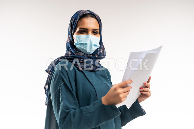 اتباع الاجراءات الاحترازية للوقاية من فايروس كورونا، بورتريه مقرب من الجانب لإمرأة عربية خلجية عمانية محجبة ترتدي العباءة وتضع الكمامة، سيدة سعودية تمسك أوراق بيضاء فارغة, الحفاظ على السلامة من كوفيد 19، لوحات توعوية لتجنب إنتشار العدوى، خلفية بيضاء