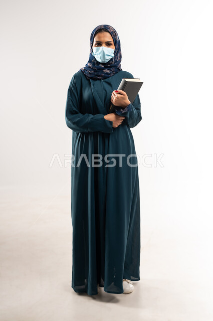 الوقوف باستقامة وثبات بايماءات الثقة، اتباع الاجراءات الاحترازية للوقاية من فايروس كورونا، بورتريه  لإمرأة عربية خلجية عمانية محجبة ترتدي العباءة وتضع الكمامة، سيدة سعودية تمسك بيديها كتاب, الحفاظ على السلامة من كوفيد 19، الطول الكامل للجسم، خلفية بيضاء