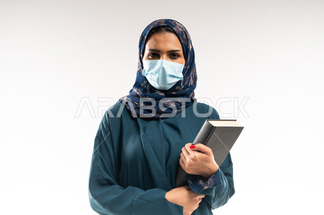 النظر إلى الكاميرا والوقوف باستقامة بايماءات الثقة، اتباع الاجراءات الاحترازية للوقاية من فايروس كورونا، بورتريه مقرب لإمرأة عربية خلجية عمانية محجبة ترتدي العباءة وتضع الكمامة، سيدة سعودية تمسك بيديها كتاب, الحفاظ على السلامة من كوفيد 19، خلفية بيضاء