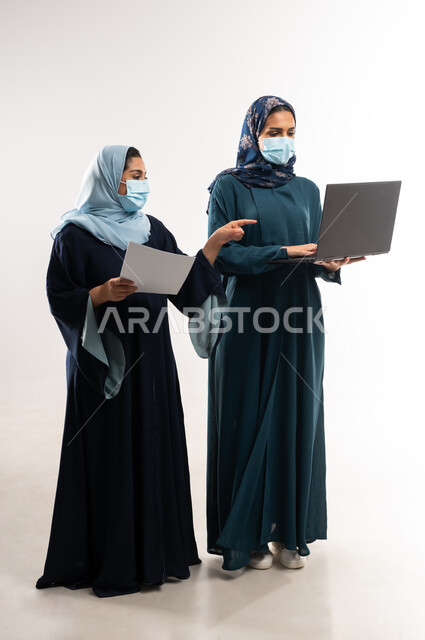 أداء المهام والعمل عن بعد، التعاون بين سيدات الأعمال السعودية في اختيار الخطة، بورتريه لامرأتين عربيتين خليجيتين عمانيتين ترتديان العباءة والكمامة تقومان بالعمل على اللابتوب، استخدام التكنولوجيا الحديثة في متابعه سير العمل، الطول الكامل للجسم، خلفية بيضاء