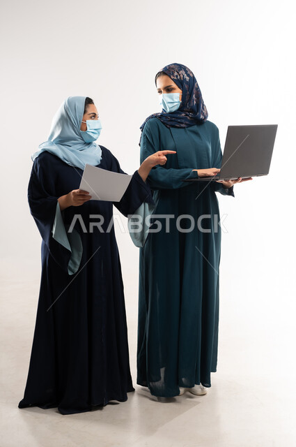 الالتزام بالتعليمات الصحية، التعاون بين سيدات الأعمال السعودية في اختيار الخطة، بورتريه لامرأتين عربيتين خليجيتين عمانيتين ترتديان العباءة والكمامة تقومان بالعمل على اللابتوب، استخدام التكنولوجيا الحديثة في متابعه سير العمل، الطول الكامل للجسم، خلفية بيضاء