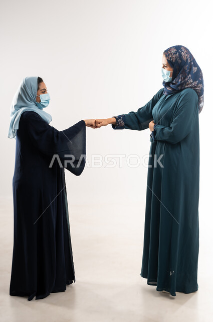 اتفاقيات عمل ناجحة لتحقيق الأهداف، الحفاظ على المسافات الآمنة وعدم التلامس، الإلتزام بتعليمات الصحة، بورتريه لامرأتين عربيتين خليجيتين عمانيتين محجبتان ترتديان العباءة والكمامة تتصافحان بقبضة اليد بايماءات السعادة، عقد صفقات بين سيدات أعمال سعودية، خلفية بيضاء