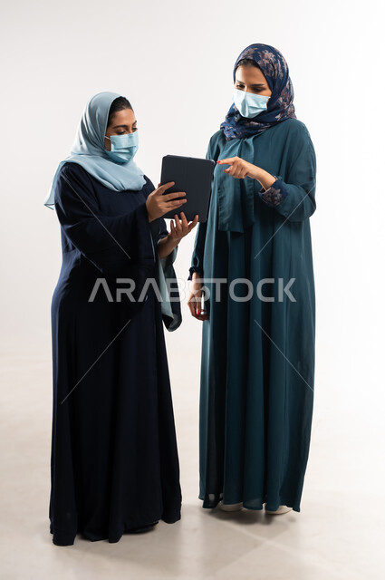 التركيز والعمل المتقن, تطوير الاعمال والشركات، استخدام التكنولوجيا الحديثة في متابعه سير العمل, التعاون بين سيدات الأعمال السعودية، بورتريه لامرأتين عربيتين خليجيتين عمانيتين ترتديان العباءة والكمامة تقومان بالعمل على جهاز التابليت، الطول الكامل للجسم، خلفية بيضاء