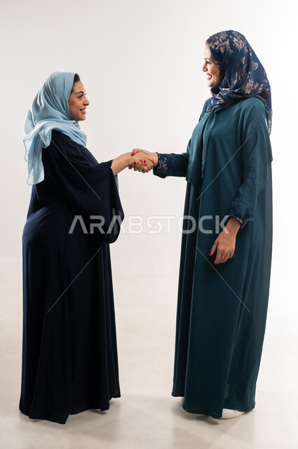 العمل في الشركات الخليجية الراقية, بورتريه لامرأتين عربيتين خليجيتين عمانيتين محجبتان ترتديان العباءة  تتصافحان بايماءات السعادة والسرور، عقد اتفاقيات وصفقات عمل ناجحة بين سيدات أعمال سعودية، تعابيرالتحية والسلام بين الزملاء, الطول الكامل للجسم، خلفية بيصاء