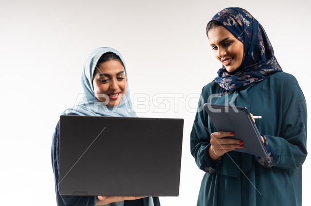 Understanding between Saudi businesswomen, close-up portrait of two Gulf Arab Omani women wearing abaya working on laptop and tablet, use of modern technology to follow up on work progress, professionalism and dedication to achievement, exchange of successful experiences, white background