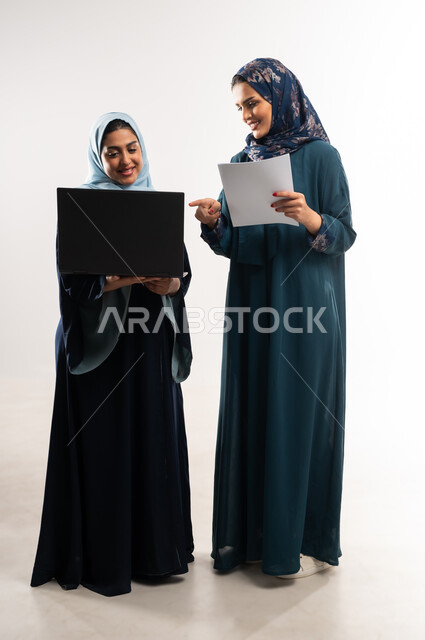 Performing tasks and working remotely, cooperation between Saudi businesswomen in choosing a business plan, portrait of two veiled Gulf Arab Omani women wearing the abaya working on a laptop, using modern technology to follow up on work progress, full body length, white background