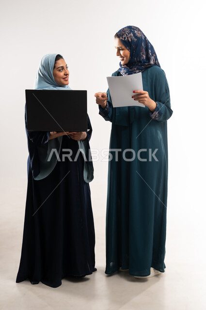 استخدام التكنولوجيا الحديثة في متابعه سير العمل، أداء المهام والعمل عن بعد، التعاون بين سيدات الأعمال السعودية في اختيار خطة العمل، بورتريه لامرأتين عربيتين خليجيتين عمانيتين محجبتان ترتديان العباءة تقومان بالعمل على اللابتوب، الطول الكامل للجسم، خلفيه بيضاء