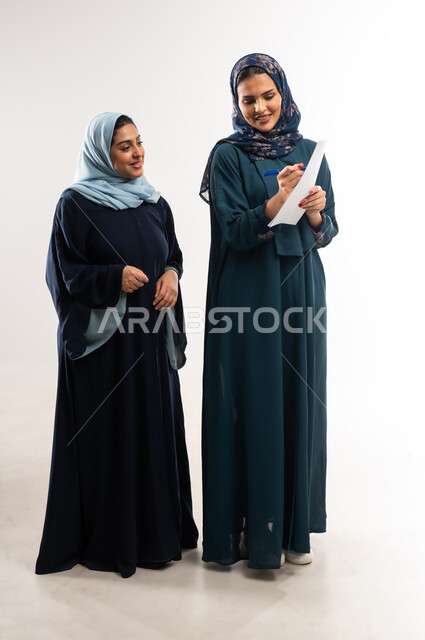 مفهوم التعاون بين سيدات الأعمال السعودية، بورتريه لامرأتينعربيتين خليجيتين عمانيتين محجبتان ترتديان العباءة تقومان بدراسة المشاريع الجديدة ومناقشتها, التشارك بين الزملاء لاختيار خطة العمل، تحقيق أهداف الشركة وحل المشكلات، الجسم بالطول الكامل, خلفية بيضاء