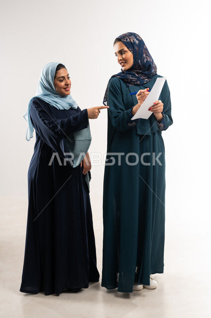 التشارك بين الزملاء لاختيار خطة العمل، تحقيق أهداف الشركة وحل المشكلات، مفهوم التعاون بين سيدات الأعمال السعودية، بورتريه لامرأتين عربيتين خليجيتين محجبتان ترتديان العباءة تقومان بدراسة المشاريع الجديدة ومناقشتها, الجسم بالطول الكامل, خلفية بيضاء