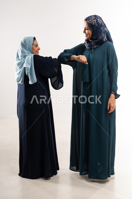 المصافحة الآمنة للإلتزام بتعليمات الصحة، بورتريه لامرأتين عربيتين خليجيتين عمانيتين ترتديان العباءة والحجاب تتصافحان بالأكواع، عقد صفقات بين سيدات أعمال سعودية، اتفاقيات عمل ناجحة لتحقيق الأهداف، الحفاظ على المسافات وعدم التلامس، الجسم بالطول الكامل، خلفية بيضاء