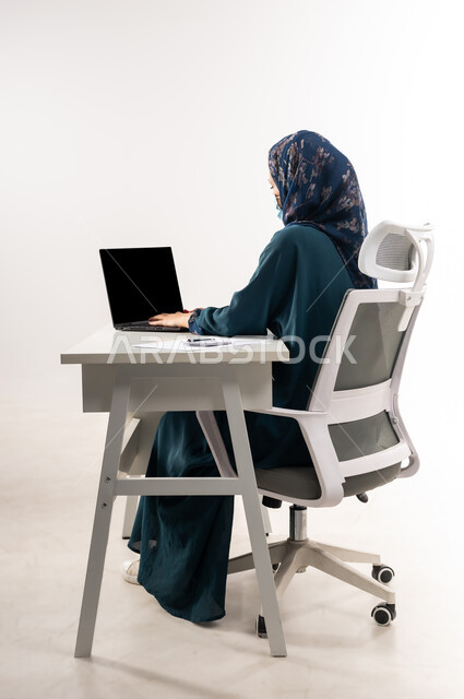 تحقيق استراتيجيات وأهداف الشركة، سيدة سعودية ترتدي الكمامة لاتخاذ الاجراءات الوقائية لفايروس كورونا, بورتريه من الجانب لإمرأة عربية خليجية عمانية ترتدي العباءة تجلس خلف المكتب وتعمل على جهاز الحاسوب المحمول, العمل عن بعد باستخدام التقنيات الحديثه، خلفية بيضاء