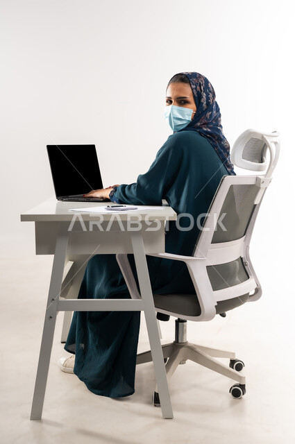 دمج التقنيات الحديثة بالحياة العملية, سيدة سعودية ترتدي الكمامة لاتخاذ الاجراءات الوقائية لفايروس كورونا, بورتريه من الجانب لإمرأة عربية خليجية عمانية ترتدي العباءة تجلس خلف المكتب وتعمل على جهاز اللابتوب، النظر إلى الكاميرا بايماءات الثقة بالنفس، خلفية بيضاء