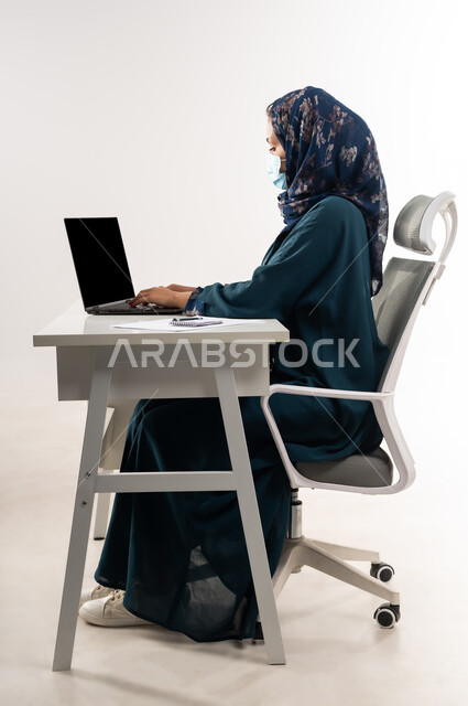 Remote work using modern technologies, achieving company strategies and goals, Saudi woman wearing a mask to take preventive measures for Corona virus, side portrait of an Arab Gulf Omani woman wearing an abaya sitting behind a desk and working on a laptop, white background