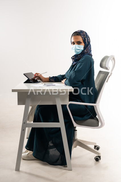 Looking at the camera with gestures of self-confidence, integrating modern technologies into practical life, a Saudi woman wearing a mask to take preventive measures for the Corona virus, a side portrait of an Arab Gulf Omani woman wearing an abaya sitting behind a desk and working on a tablet, white background