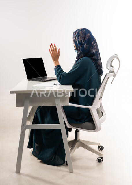 استخدام الاجهزة والتقنيات الحديثة, بورتريه لإمرأة عربية خليجية عمانية ترتدي العباءة تجلس خلف المكتب تقوم باجراء مكالمة فيديو عبر جهاز اللابتوب بايماءات القاء التحية والسلام، سيدة سعودية تضع الكمامة الطبية لاتباع الاجراءات الوقائية ضد فايروس كورونا، خلفية بيضاء