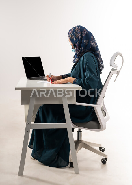 مفهوم الاجتهاد وانجاز الاعمال المطلوبة، عرض شاشة سوداء فارغة على جهاز اللابتوب، بورتريه من الجانب لإمرأة عربية خليجية عمانية ترتدي العباءة تجلس خلف المكتب تقوم بتدوين الملاحظات, سيدة سعودية تضع الكمامة لاتخاذ الاجراءات الوقائية لفايروس كورونا, خلفية بيضاء