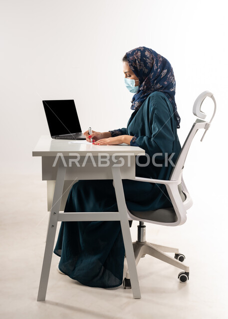 عرض شاشة سوداء فارغة على جهاز اللابتوب، بورتريه من الجانب لإمرأة عربية خليجية عمانية ترتدي العباءة تجلس خلف المكتب تقوم بتدوين الملاحظات, سيدة سعودية تضع الكمامة لاتخاذ الاجراءات الوقائية لفايروس كورونا, مفهوم الاجتهاد وانجاز الاعمال المطلوبة، خلفية بيضاء