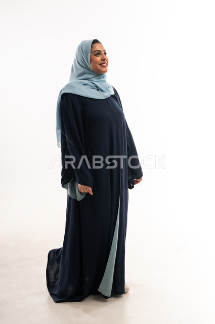 مفهوم الأناقة والأنوثة, بورتريه لامرأة عربيه عمانية محجبة ترتدي العباءه تنظر الى شيء ما, سيدة سعودية تقف باستقامة واعتدال بايماءات الثقة بالنفس, النظرة المستقبلية والتفكير لتحقيق النجاح, الاهتمام بالمظهر الخارجي، الطول الكامل للجسم, خلفية بيضاء