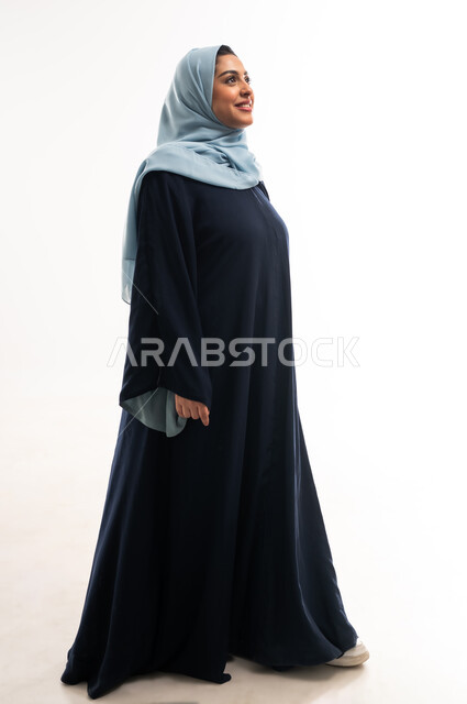 التقدم إلى الأمام بخطوات ثابتة، سيدة سعودية تمشي بتعابير السعادة، الاهتمام بالمظهر الخارجي، مفهوم الأناقة والأنوثة, ايماءات الذهاب إلى مكان معين، بورتريه من الجانب لإمرأة عربية خليجية عمانية محجبة ترتدي العباءة تنظر إلى شيء ما، الطول الكامل للجسم، خلفية بيضاء