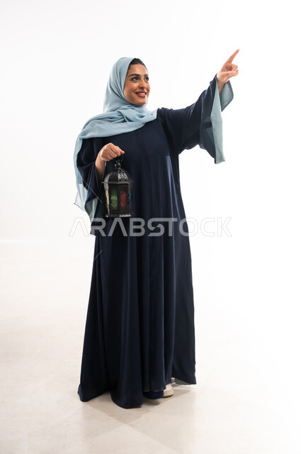 الإستعداد لقدوم شهر رمضان المبارك، المناسبات والأعياد الدينية، العبادة والتقرب إلى الله، بورتريه لإمراة عربية خليجية عمانية محجبة ترتدي العباءة تحمل بيدها فانوس معدني، سيدة سعودية تشير بإصبع السبابة الى شيء ما، الطول الكامل للجسم، خلفية بيضاء