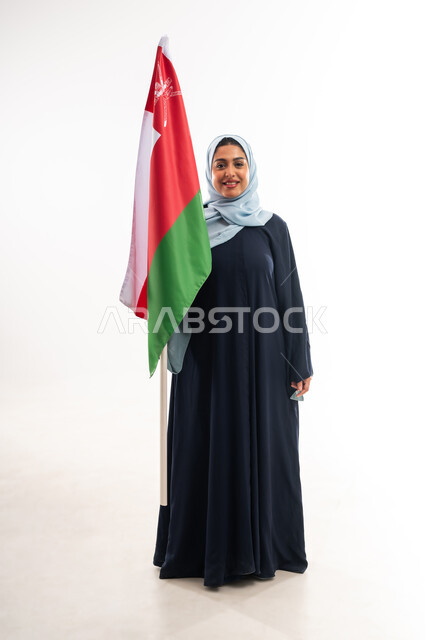 Participating in national holidays and occasions, celebrating the Omani National Day on November 18, gestures of pride and honor, portrait of a veiled young Omani Gulf Arab woman wearing an abaya holding the flag of the Sultanate of Oman, the concept of love of the homeland and belonging to it, full-length body, white background