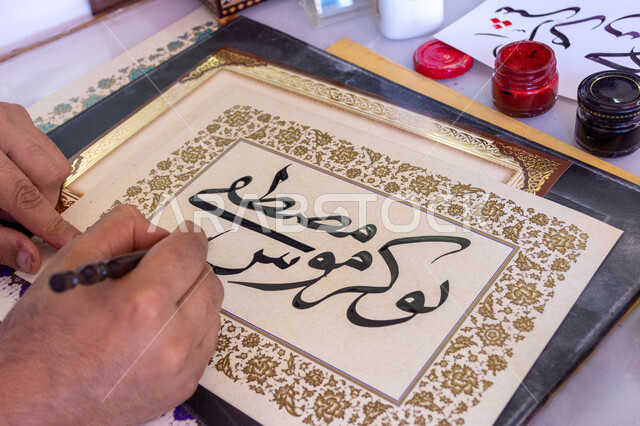 احتراف الكتابة بخط الثلث، فنون الخط العربي، صورة مقربة ليد رجل خطاط عربي خليجي سعودي يمسك بيده الريشة الخاصة ويكتب على لوحة أمامه، تعليم مهارات الرسم بالخطوط بشكل انسيابي، التمسك بالثقافة العربية الأصيلة في السعودية، نقش الكلمات وكتابتها بشكل فني هندسي مميز