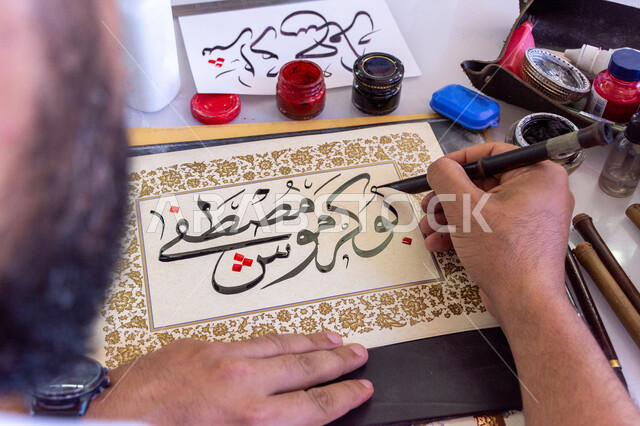 فنون الخط العربي، احتراف الكتابة بخط الثلث، صورة مقربة ليد رجل خطاط عربي خليجي سعودي يمسك بيده الريشة الخاصة ويكتب على لوحة أمامه، تعليم مهارات الرسم بالخطوط بشكل انسيابي، التمسك بالثقافة العربية الأصيلة في السعودية، نقش الكلمات وكتابتها بشكل فني هندسي مميز
