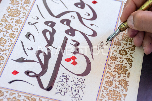 احتراف الكتابة بخط الثلث، فنون الخط العربي، صورة مقربة ليد رجل خطاط عربي خليجي سعودي يمسك بيده الريشة الخاصة ويكتب على لوحة أمامه، تعليم مهارات الرسم بالخطوط بشكل انسيابي، التمسك بالثقافة العربية الأصيلة في السعودية، نقش الكلمات وكتابتها بشكل فني هندسي مميز