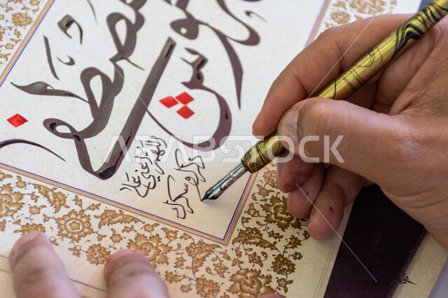 فنون الخط العربي، احتراف الكتابة بخط الثلث، صورة مقربة ليد رجل خطاط عربي خليجي سعودي يمسك بيده الريشة الخاصة ويكتب على لوحة أمامه، تعليم مهارات الرسم بالخطوط بشكل انسيابي، التمسك بالثقافة العربية الأصيلة في السعودية، نقش الكلمات وكتابتها بشكل فني هندسي مميز