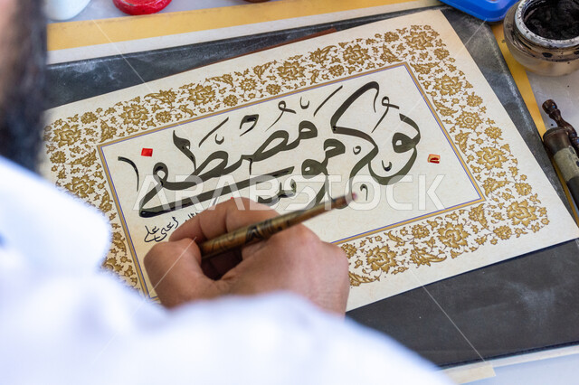 فنون الخط العربي، احتراف الكتابة بخط الثلث، صورة مقربة ليد رجل خطاط عربي خليجي سعودي يمسك بيده الريشة الخاصة ويكتب على لوحة أمامه، تعليم مهارات الرسم بالخطوط بشكل انسيابي، التمسك بالثقافة العربية الأصيلة في السعودية، نقش الكلمات وكتابتها بشكل فني هندسي مميز
